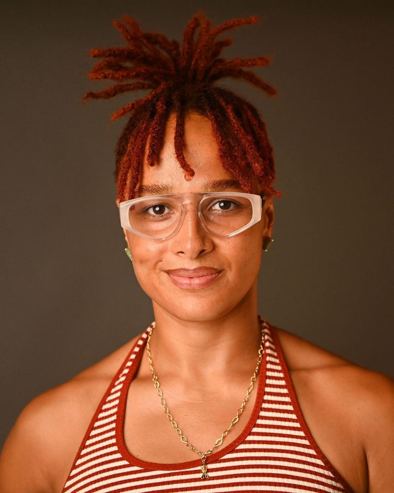 Portrait of Balla-Gabriele, with red locs tied up, clear-framed glasses and a red and white striped top.